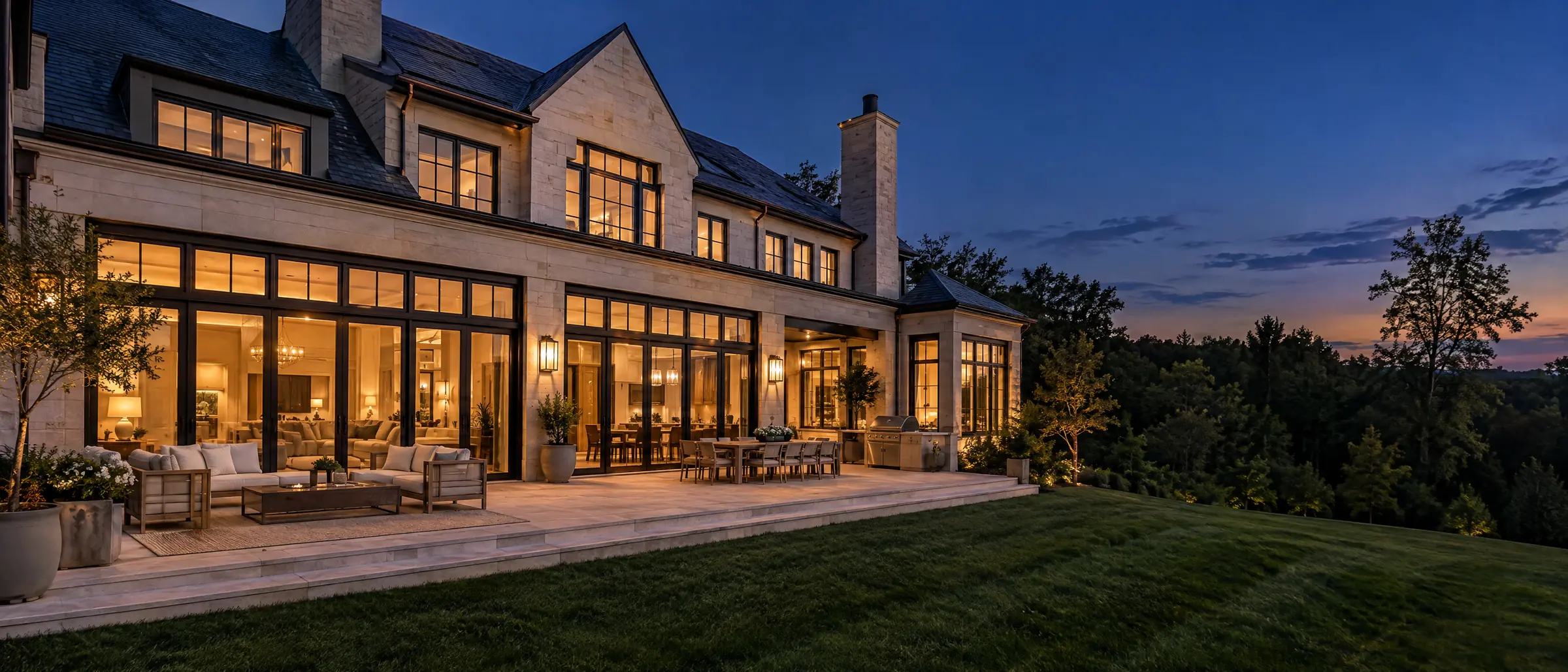 The back of a finished custom home at twilight, a stone terrace before a wall of lit glass doors