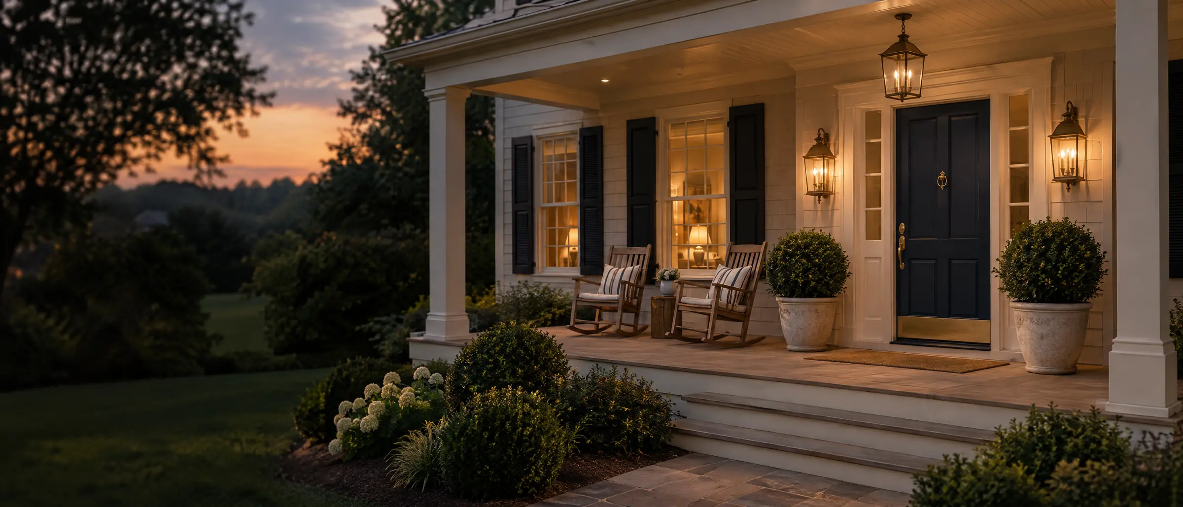 A covered front porch at dusk with a navy door, brass lanterns, and two rocking chairs