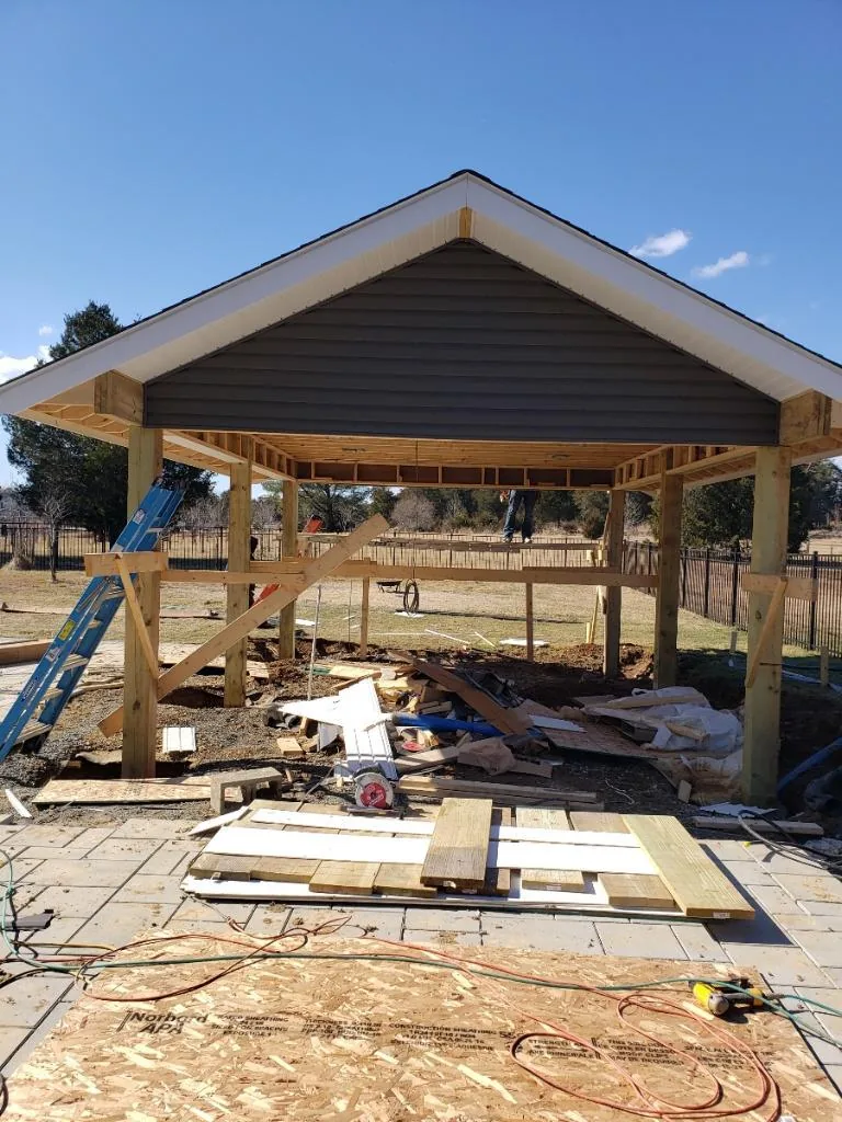 A new covered porch under construction, its timber posts and gable roof framing complete against a blue sky.
